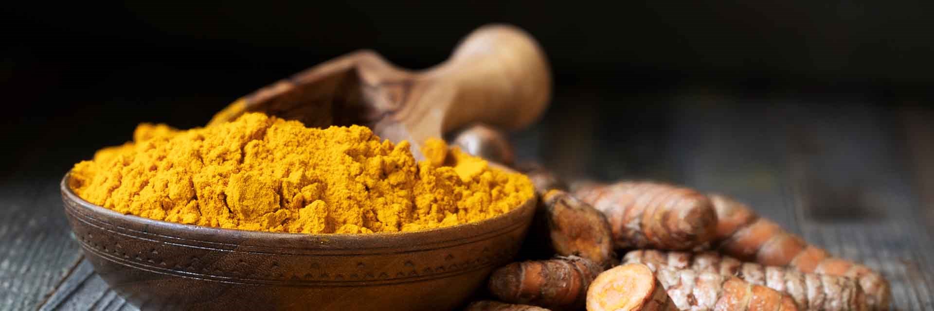 A wooden bowl of the bright orange turmeric powder, with fresh turmeric next to the bowl