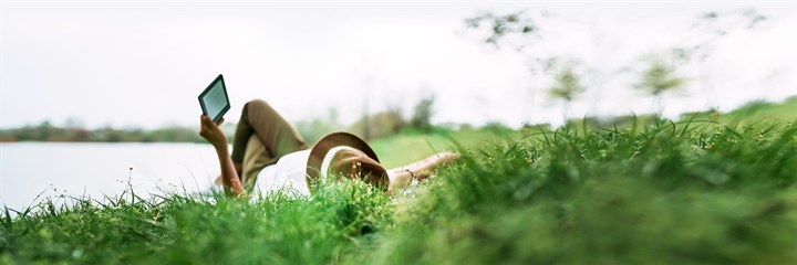 Man lying in grass by river reading ebook