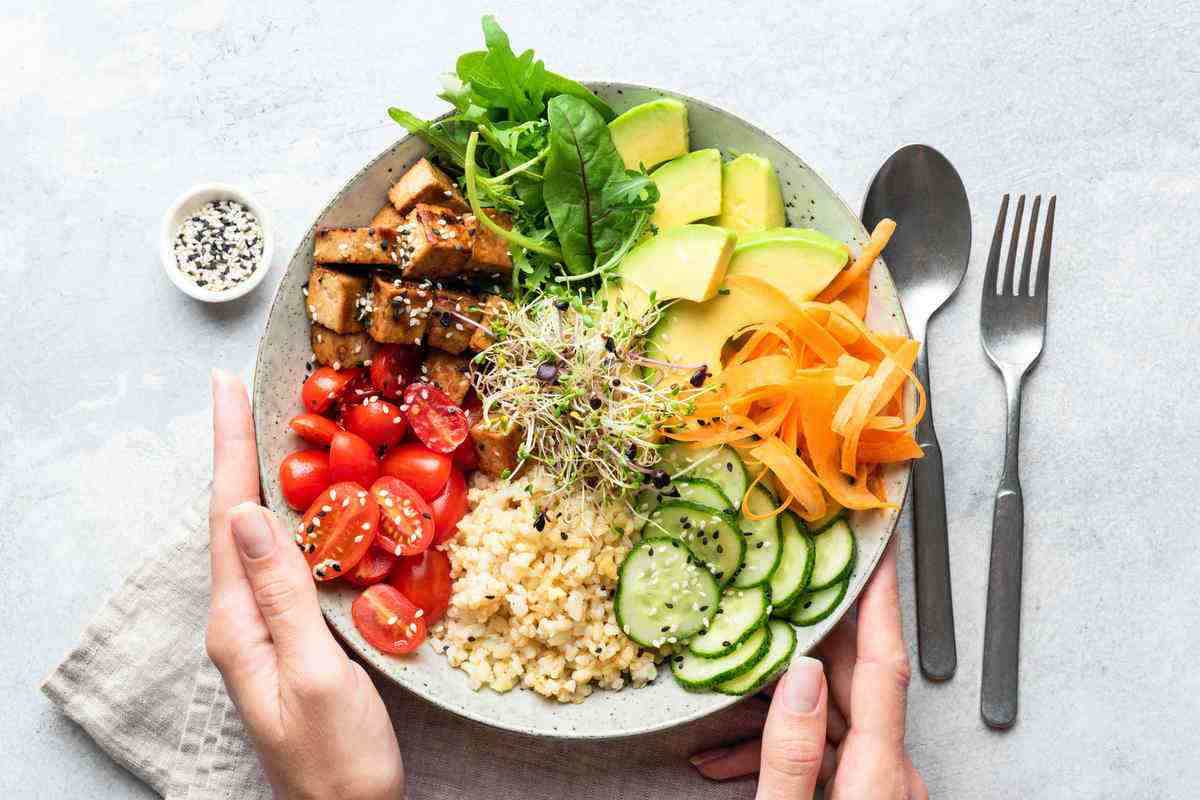 Buddha bowl salad with tofu, avocado and other vegetables in female hands, balanced meal. Bright grey concrete background, table top view