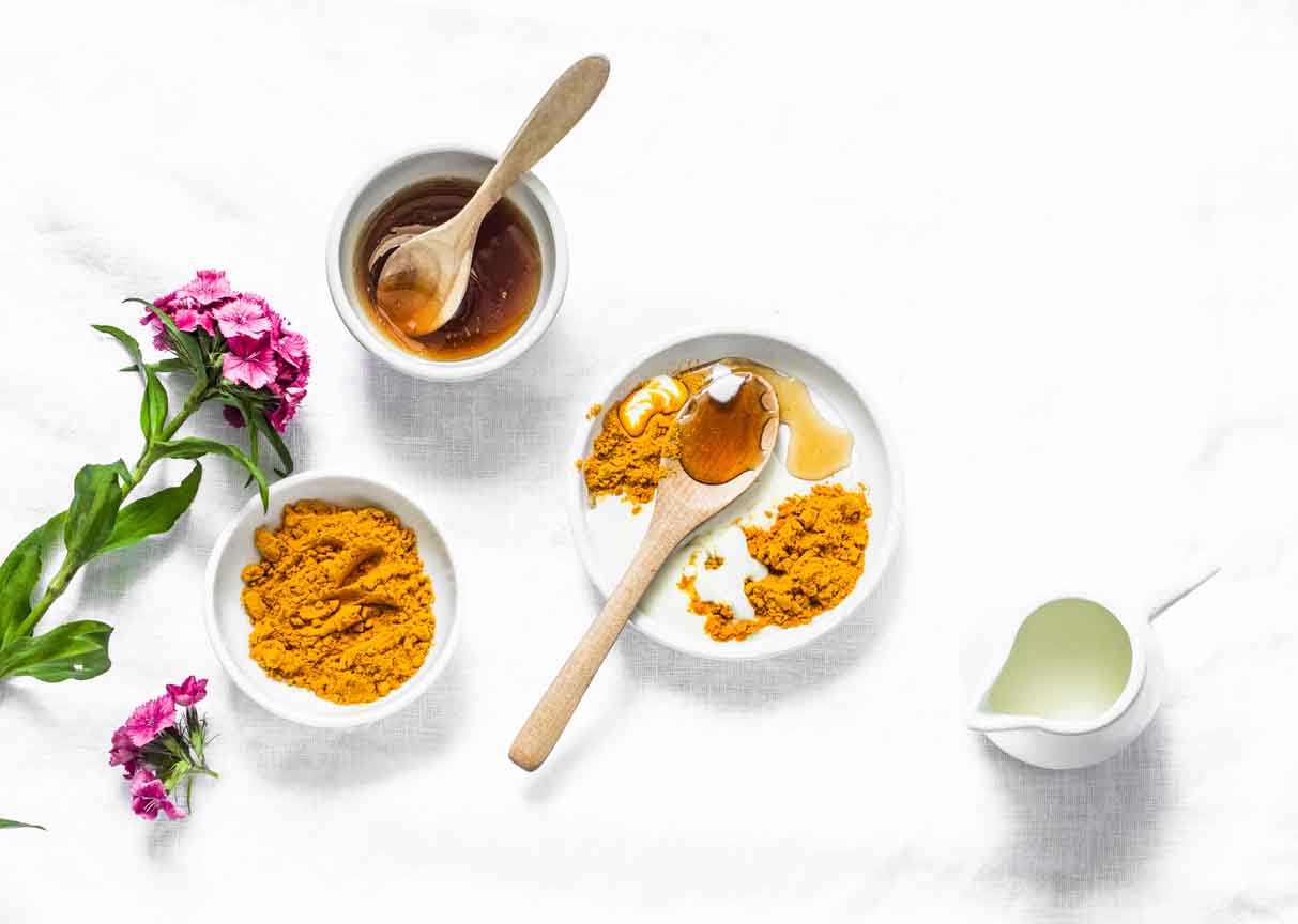 Overhead photo of a dish of turmeric powder, a jug of coconut milk, and a bowl of honey, with wooden spoons