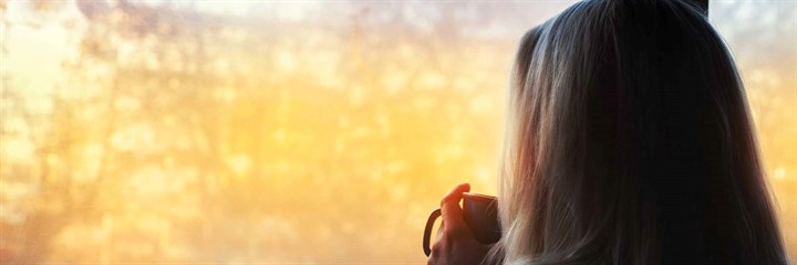Grey-haired woman holding mug looking out window at sunset