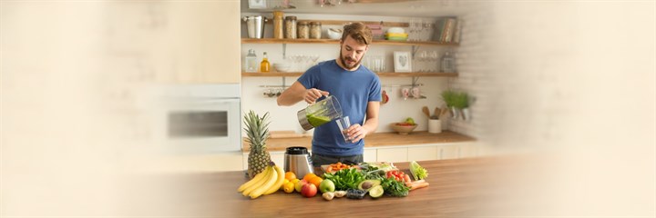 Man in kitchen pouring green juice from a blender into a glass, surrounded by fruit and vegetables
